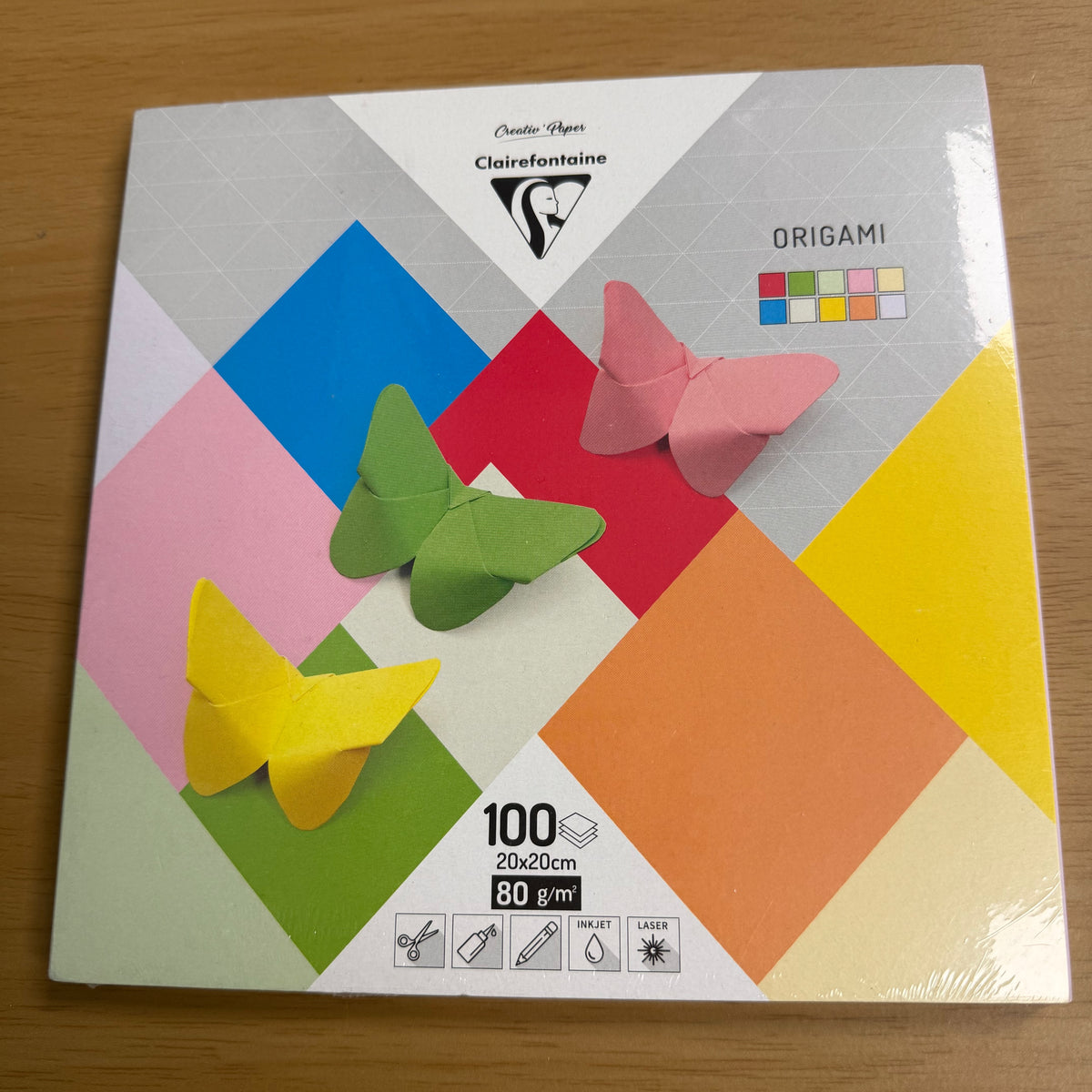 The retail box for the product. It features a pink, red, white, yellow, green and blue diamond design and displays three images of origami butterflies. One in pink, one in green and one in yellow.