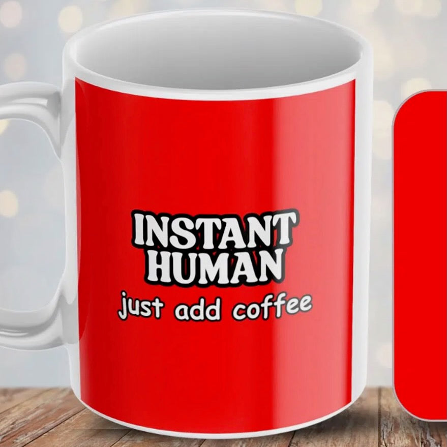 A ceramic mug with a red wraparound print with the words "Instant Human just add coffee' in white text with a black outline.