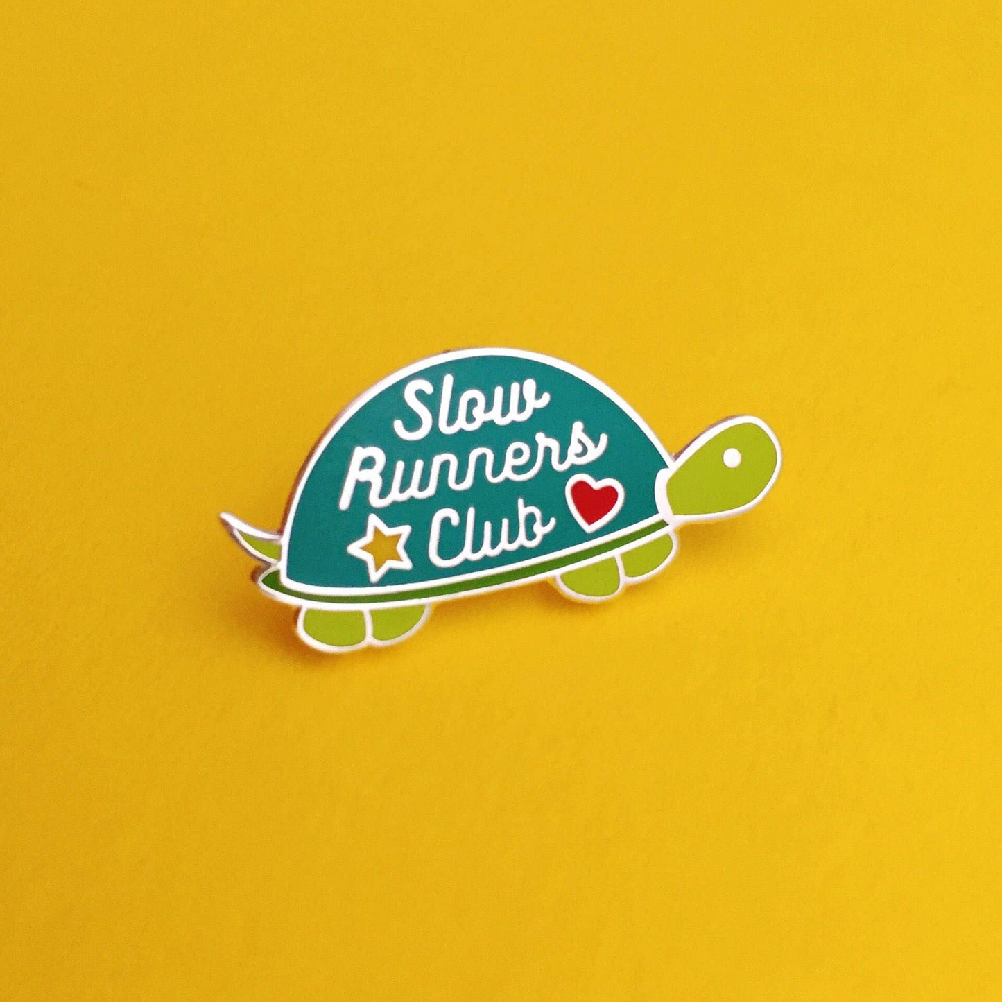 A turtle-shaped enamel pin with a dark dreen shell, silver border, green head and legs, and the words 'Slow Runners Club' with a small red heart and yellow star, shown on a solid yellow background.
