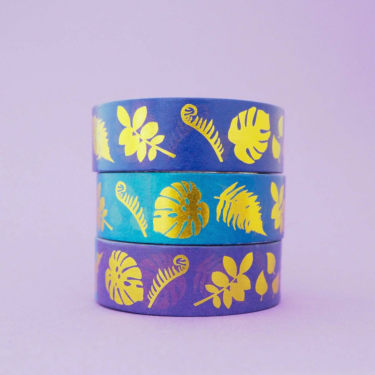 Three rolls of washi tape stacked vertically, featuring tropical leaf and fern patterns in blue, turquoise, and purple with metallic gold foil accents, set against a solid light purple background.