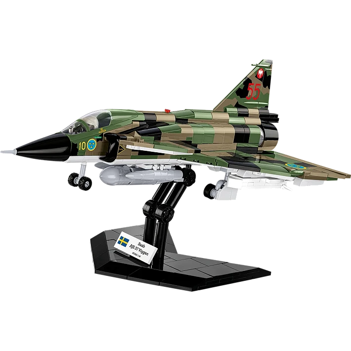 A photo of the assembled model plane, stood on its black display stand. The jet consists of various green, brown and black blocks, features a black nose cone, transparent cockpit wind shield. and has several grey and white bombs and missiles mounted beneath it. A small white plaque displaying the aircraft’s name and country of origin sits on the display stand. 