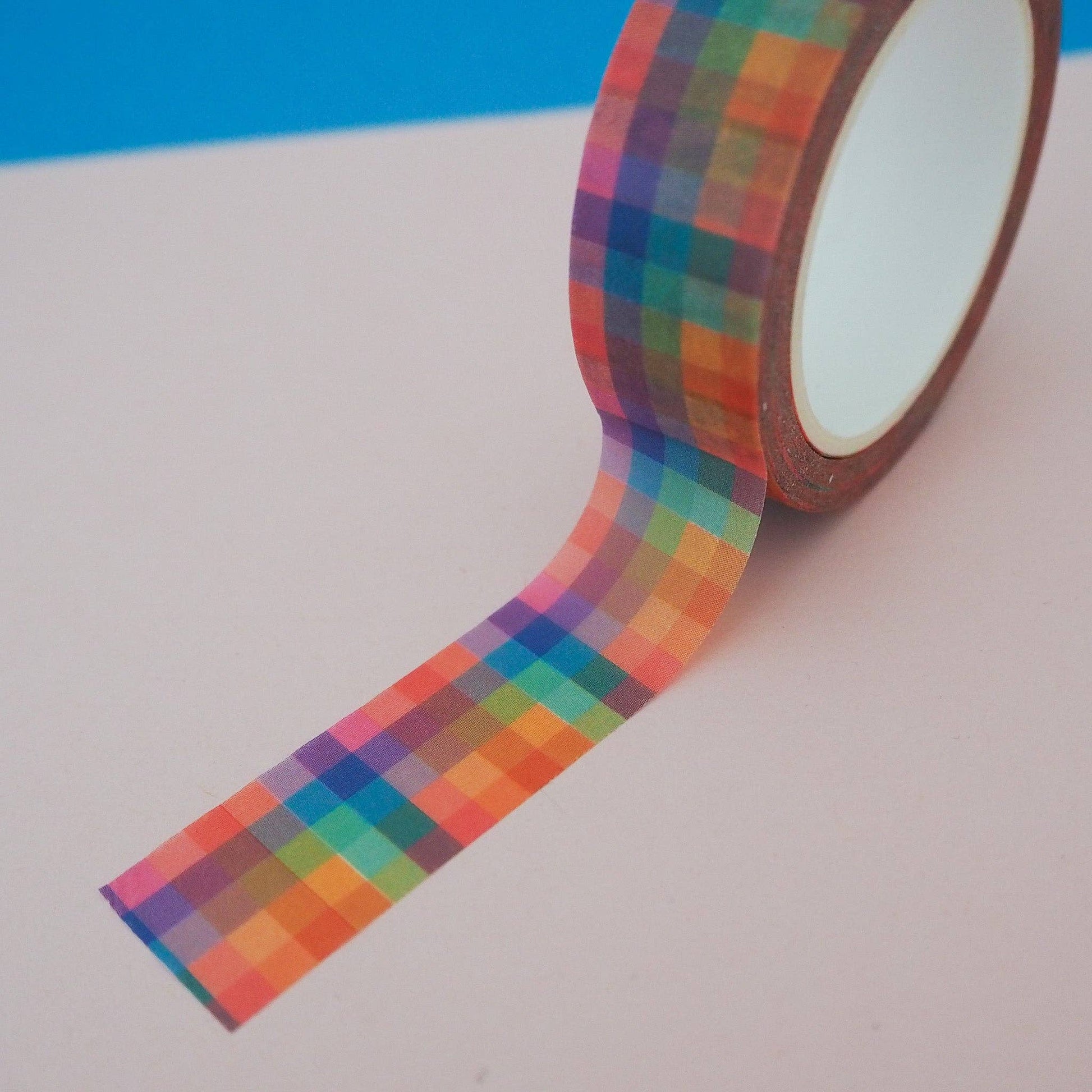Washi tape with a rainbow check pattern, partially unrolled, on a cream and blue background.