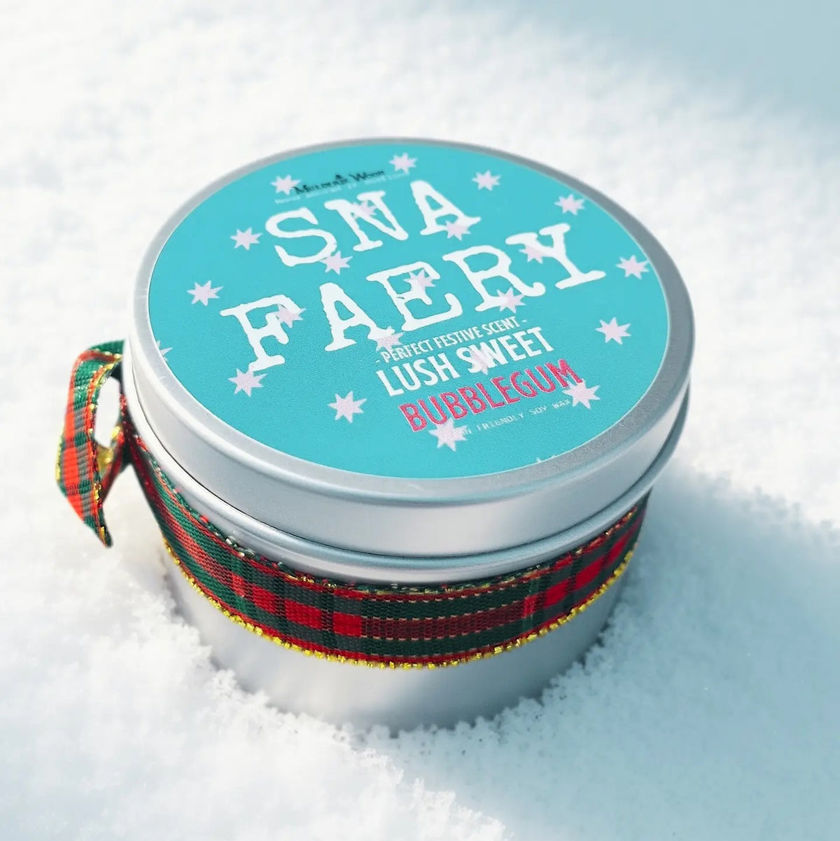 A round silver tin containing 'Sna Faery' candle sits in snow, wrapped with a red and green plaid ribbon. The lid features a bright turquoise label with white text and star graphics.