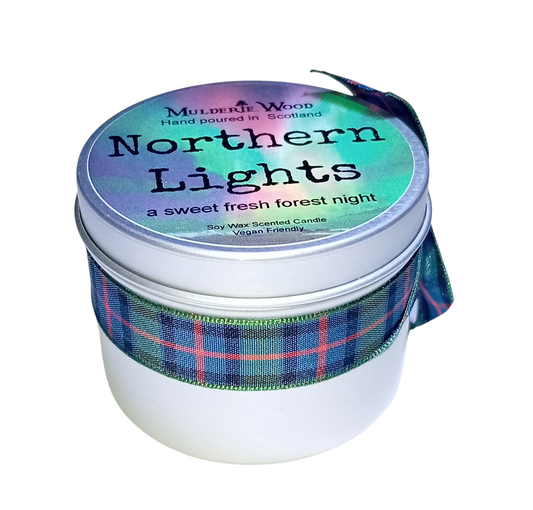 The tin the candle comes in. It has an image of the green, blue and red aurora on the top, with the text "NORTHERN LIGHTS a sweet fresh forest night" written on it in black, as well as the logo of the maker, and a small piece of text saying "Soy wax scented candle. Vegan Friendly." It has a plaid pattern green, blue and red ribbon around it.