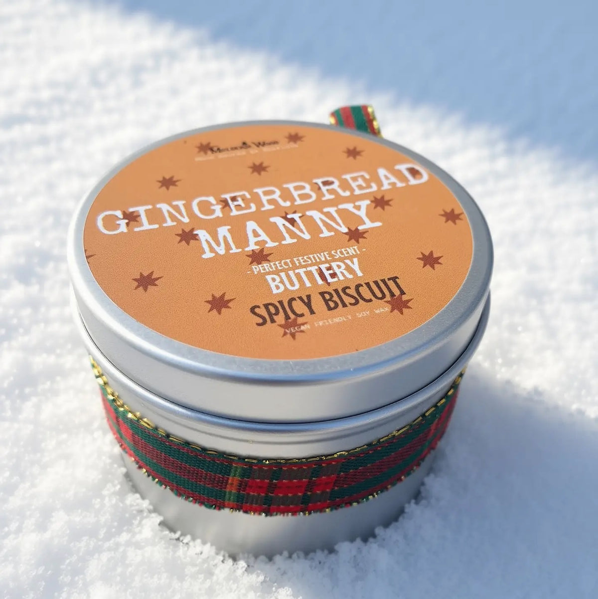 A round metal tin containing Gingerbread Manny Spicy Biscuit soy wax candle sits in the snow, wrapped in a red and green plaid ribbon with a matching round orange label on top.