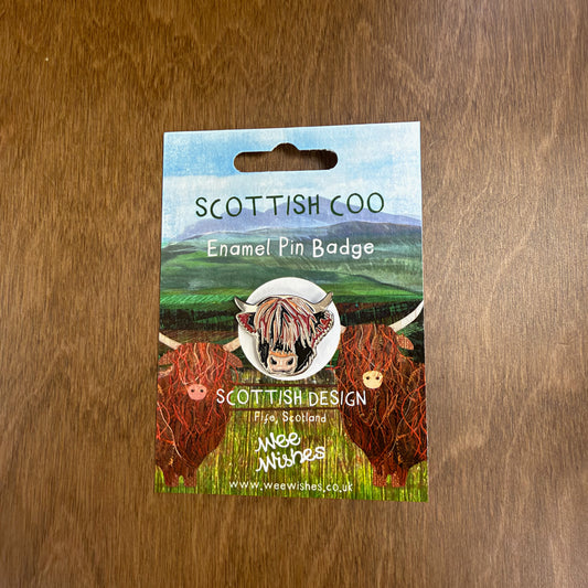 An enamel pin featuring a Highland Cow head, on a cardboard back with an illustration of highland cows in a field with mountains in the background.