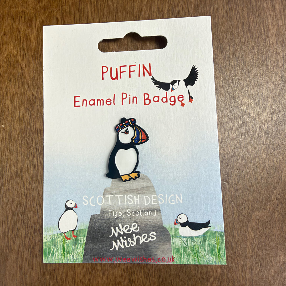 An emamel pin featuring a puffin wearing a tam o'shanter, on a iece of card with other puffins on.