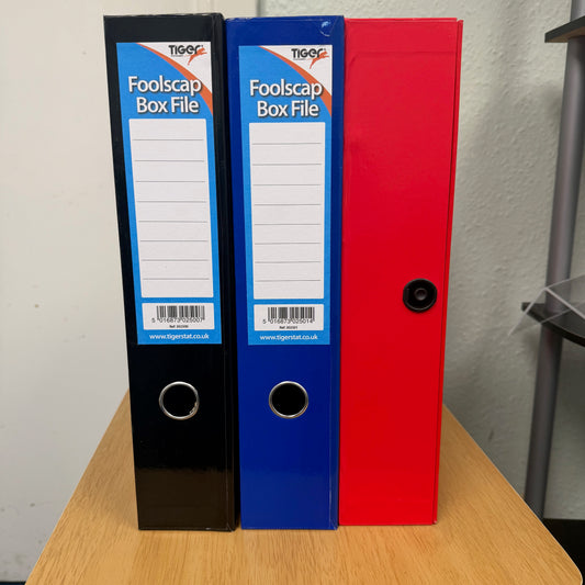 Three Foolscap box files in black, blue, and red standing upright on a wooden table against a light wall. Two files have white labels attached with RTiger branding, the third shows the spring closure for the lid.