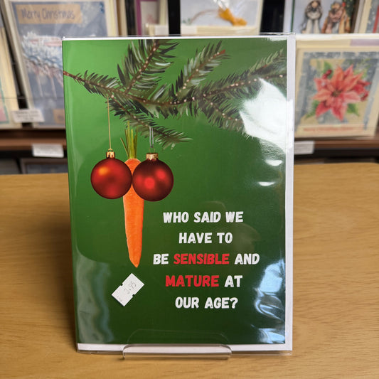 A photo of the card on a table top, with other greeting cards behind.
A green greeting card with a festive holiday illustration, featuring pine branches, red ornaments, and a carrot with the text 'WHO SAID WE HAVE TO BE SENSIBLE AND MATURE AT OUR AGE?' on a white background.
The baubles and carrot are hung in such a way as to mimic male genitalia.