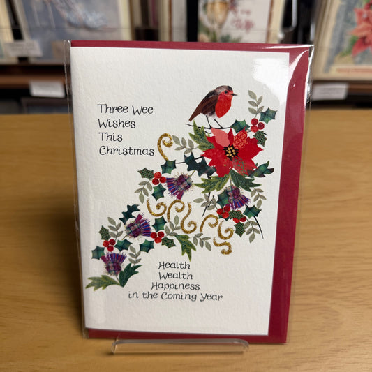 A Christmas greeting card in clear plastic packaging, standing on a small clear display. The card has a white front with a red border, a robin bird, holly leaves, pinecones, and festive greenery, plus the text 'Three Wee Wishes This Christmas' and 'Health Wealth Happiness in the Coming Year'.