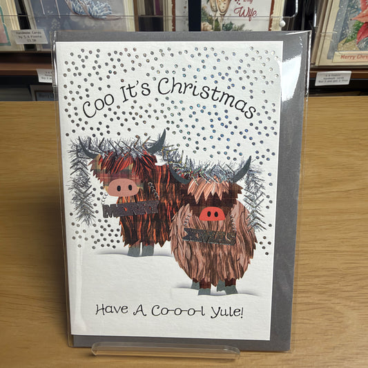A Christmas greeting card displayed on a wooden shelf. The card features two shaggy Highland cows with red snouts and black horns, set against a white background with silver glittery snowflakes and the text 'Coo It's Christmas' and 'Have A Coo-ol Yule!'.