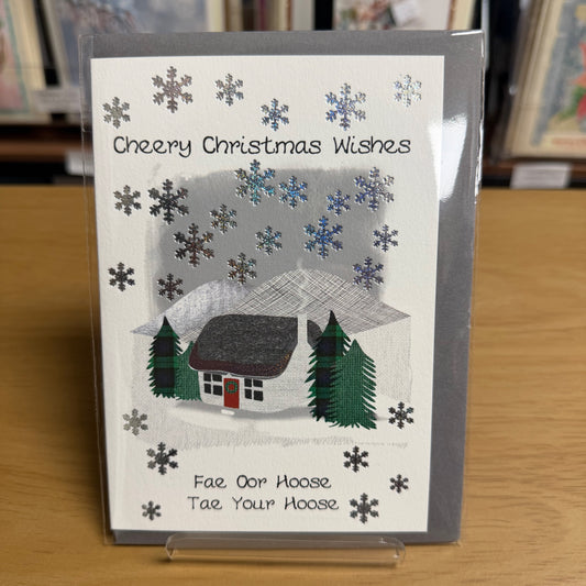 A Christmas greeting card in a clear plastic sleeve, featuring a snowy winter scene with a small house, trees, and silver snowflakes. The text reads 'Cheery Christmas Wishes' at the top and 'Fae Our Hoose Tae Your Hoose' at the bottom.