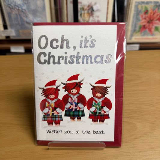 A greeting card standing on a clear display easel on a wooden table. The card has a white center with a dark red border, the text 'Och, it's Christmas' in gray, and an illustration of three red Scottish Highland cows in red suits and tartan kilts.