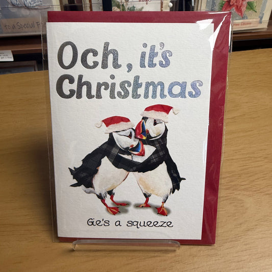 A Christmas greeting card displayed on a clear stand on a wooden table. The card has a red border, the text 'Och, it's Christmas' and 'Gie's a squeeze', and an illustration of two puffins hugging wearing Santa hats.