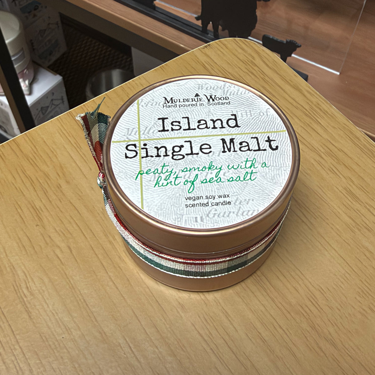 A round tin candle labeled 'Island Single Malt' by Mulderie Wood, wrapped in a green, red, and white patterned ribbon, sitting on a light wood tabletop.