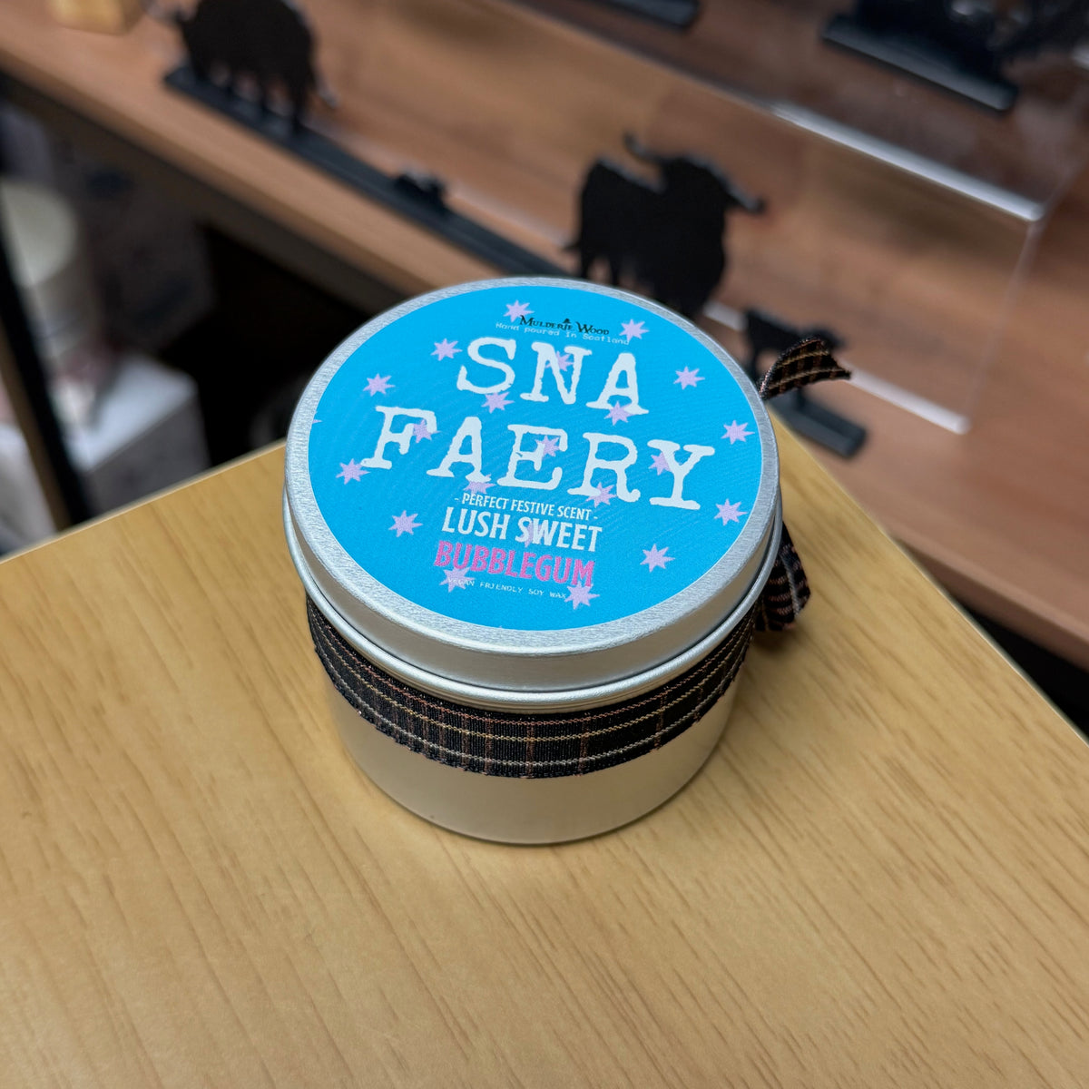 A round metal tin of Sna Faery bubblegum soy wax candle, featuring a light blue lid with white text and a dark plaid ribbon, resting on a light wooden table.