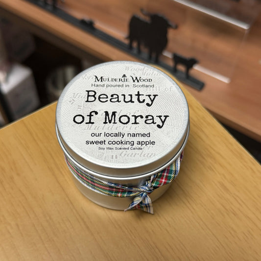 A round metal tin candle labeled 'Beauty of Moray' by Muldarie Wood, tied with a plaid ribbon, sitting on a wooden tabletop.