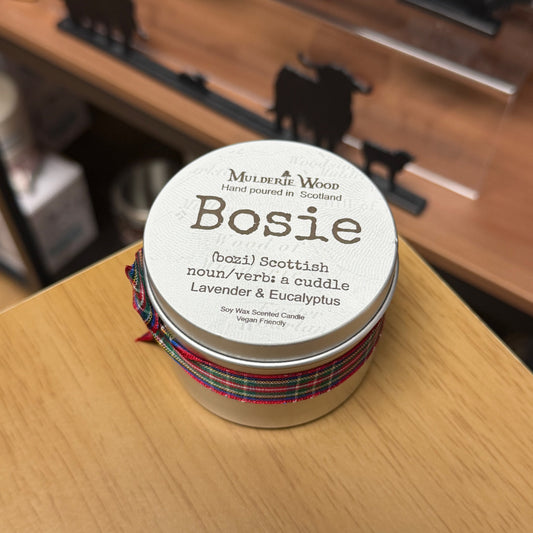A round metal tin candle labeled 'Bosie' by Mulderie Wood, wrapped with a red, green, and blue tartan ribbon, sitting on a light wood tabletop.