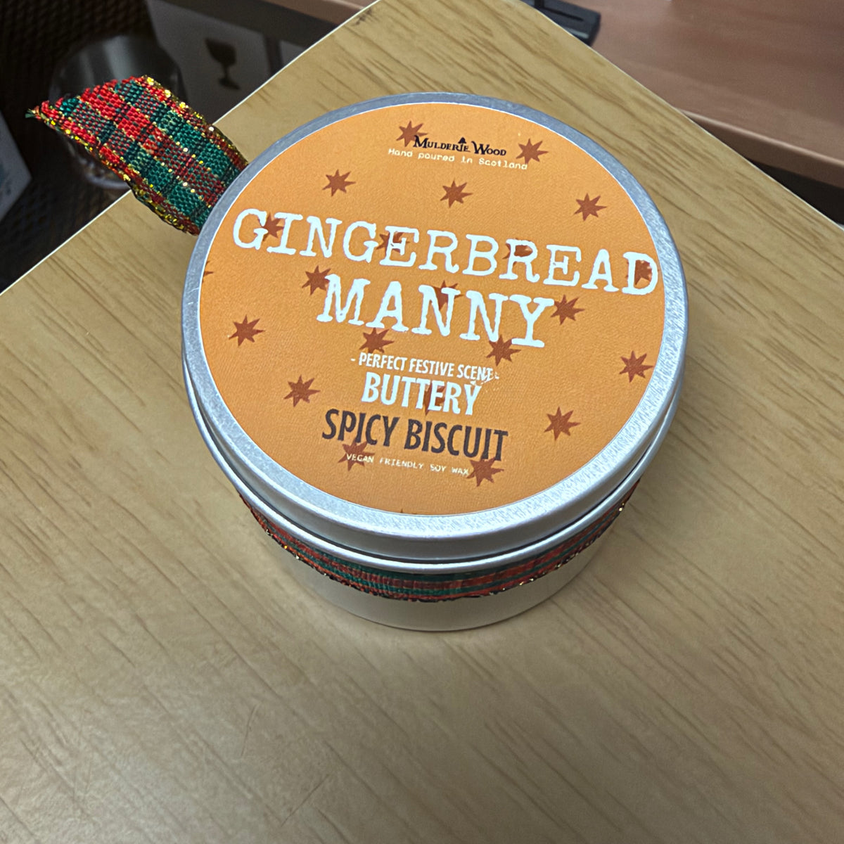 A round metal tin of 'Gingerbread Manny' scented candle by Muldarie Wood sits on a light wood tabletop. The lid features a warm orange label with white and brown text, and the tin is wrapped in a red, green and blue patterned ribbon.