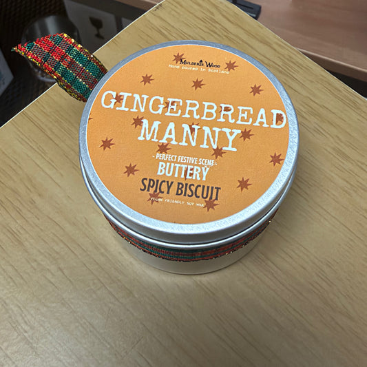 A round metal tin of 'Gingerbread Manny' scented candle by Muldarie Wood sits on a light wood tabletop. The lid features a warm orange label with white and brown text, and the tin is wrapped in a red, green and blue patterned ribbon.