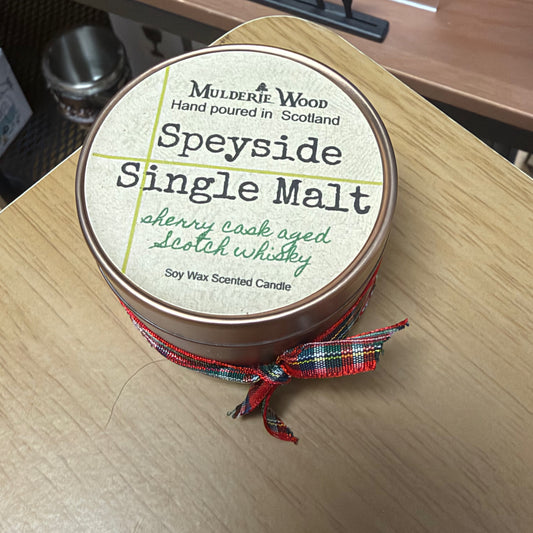 A round tin candle labeled 'Speyside Single Malt' by Mulderie Wood, tied with a red tartan ribbon, photographed on a light wood tabletop.