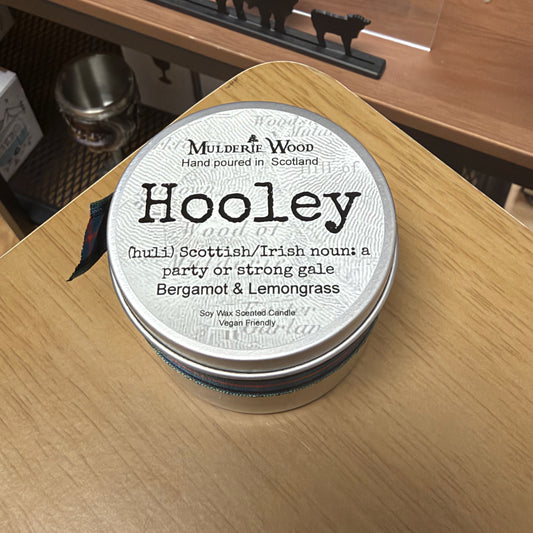 A round metal tin candle labeled 'Hooley' by Mulderie Wood, placed on a light wood tabletop. The lid is white with black text.
