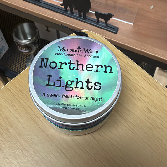 A round metal tin of Mulderie Wood 'Northern Lights' soy wax scented candle sits on a light wood tabletop. The lid features an iridescent pastel label with black text reading 'Northern Lights' and 'a sweet fresh forest night'.