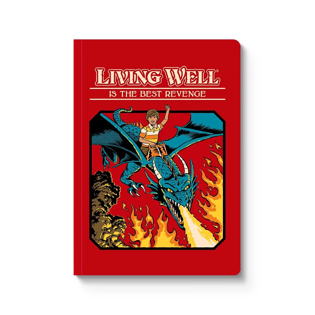 A red hardcover notebook standing upright on a white background. The cover features the title 'LIVING WELL IS THE BEST REVENGE' in stylised white and yellow lettering above an illustration of a blue dragon breathing fire, with a small figure riding it.