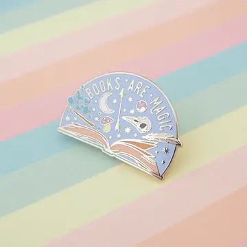 A pastel, semicircular enamel pin with an open book under a crescent moon and stars, with the phrase 'BOOKS ARE MAGIC' in silver lettering. The pin is photographed on a striped pastel background.