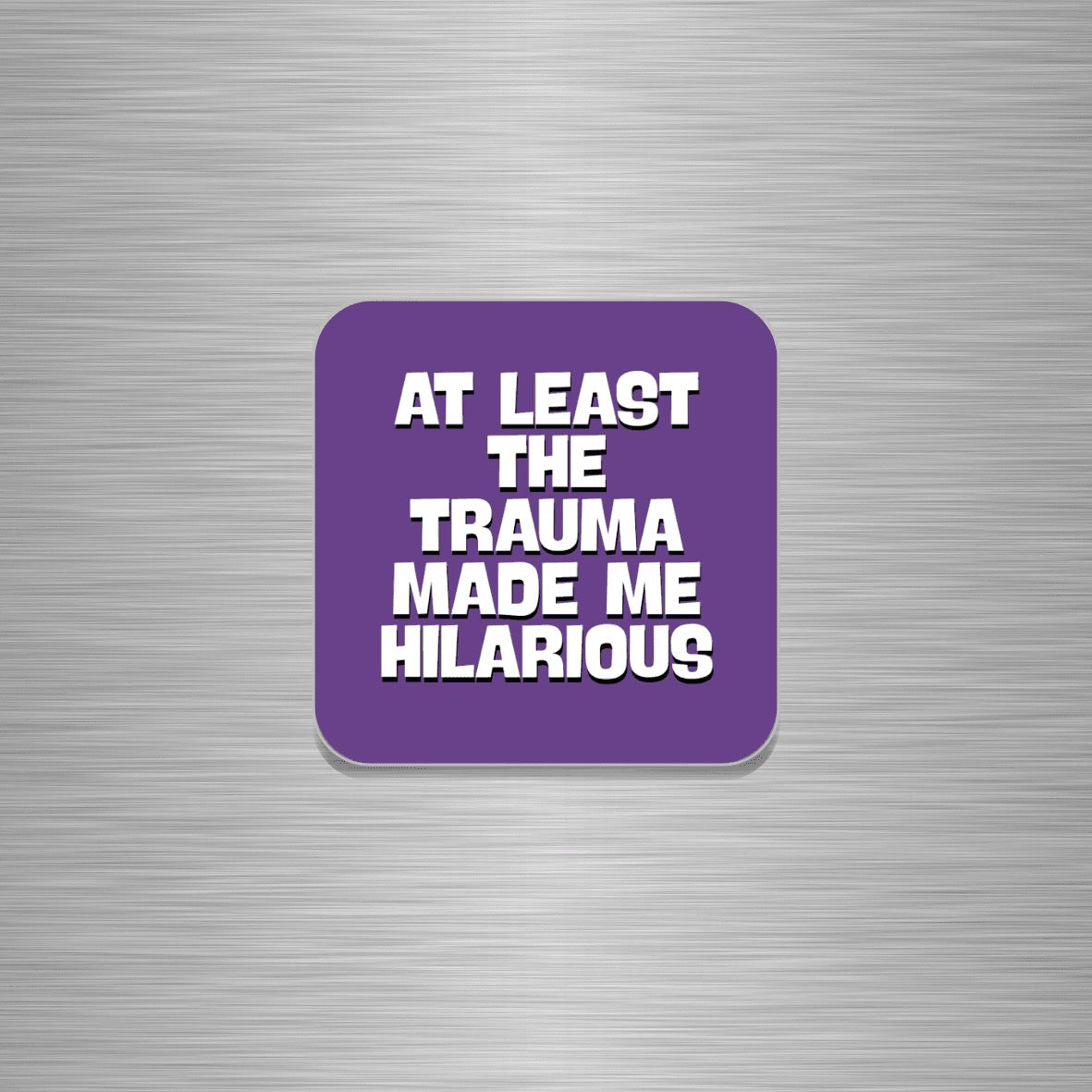 A purple rounded square fridge magnet with bold white text reading 'AT LEAST THE TRAUMA MADE ME HILARIOUS' centred on a brushed silver metal background.