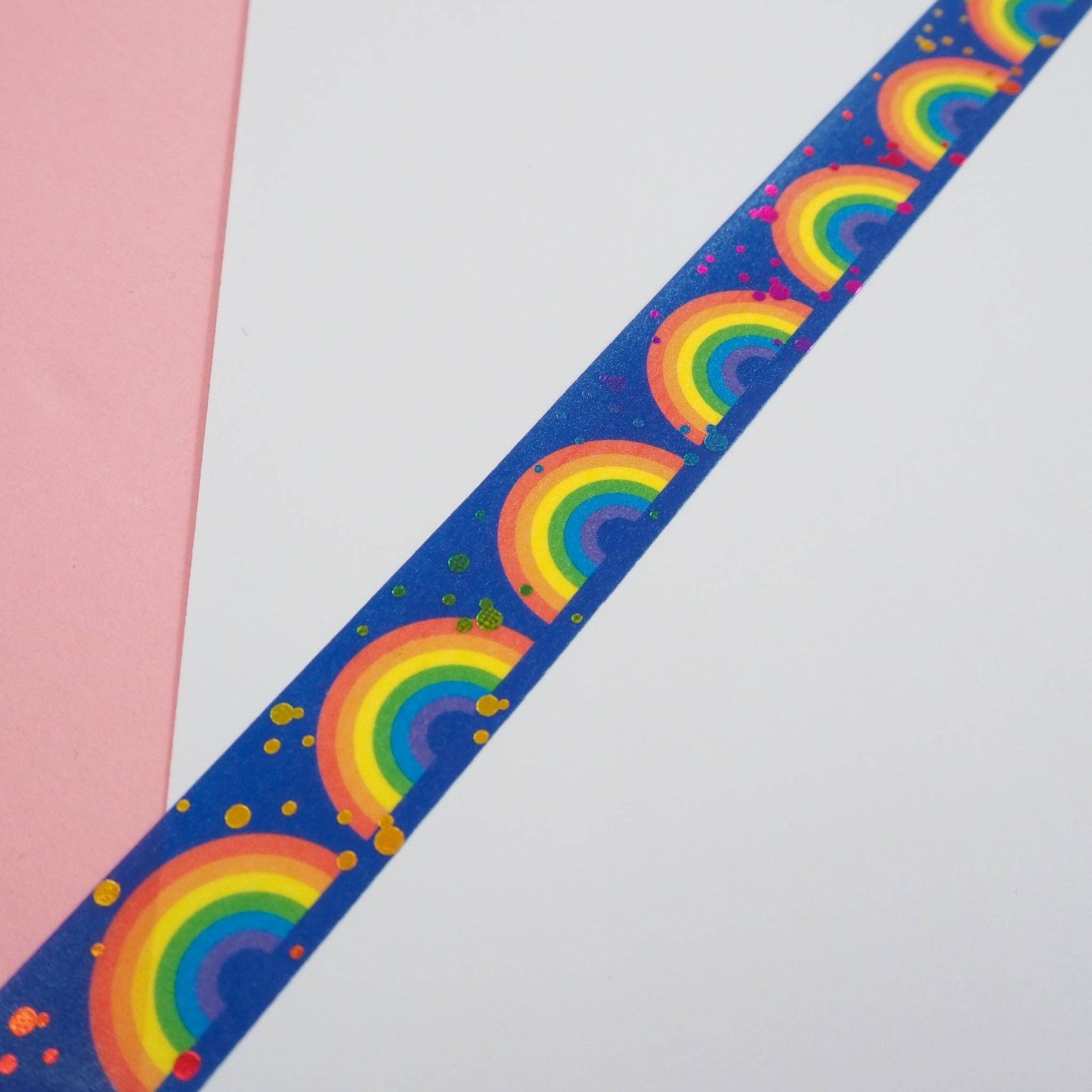 Washi tape unrolled, with a repeating pattern of rainbows and multicoloured dots, on a white piece of paper and a pink background.