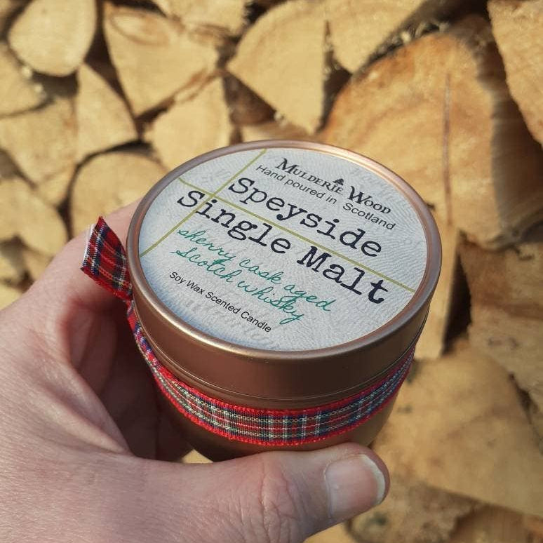 A hand holds a round tin candle in front of a stacked woodpile. The tin has a copper-toned metal body, a white label reading 'Muldarie Wood Speyside Single Malt Scotch Whisky Candle,' and a red tartan ribbon around the lower portion.