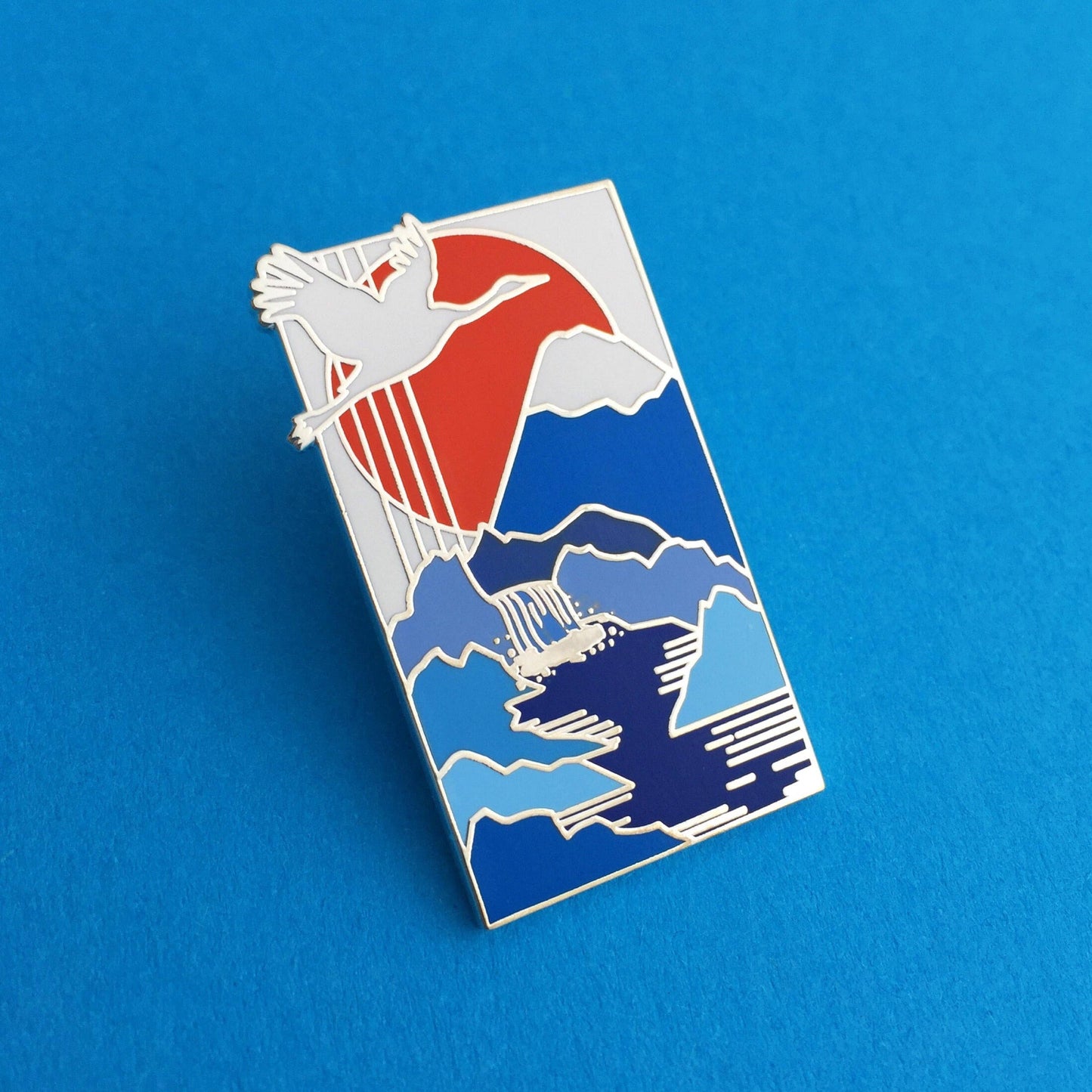 A rectangular pin, with an image of a Japanese style painting of a white crane flying across a red sun, over a snow capped mountains with a river flowing from the mountain.
Pin is on a blue background.