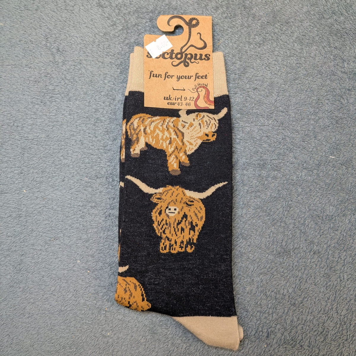 A pair of black novelty socks featuring tan illustrations of highland cows, with tan cuffs and heels. The socks are displayed on a textured grey background with their cardboard hanger tag attached to the top.