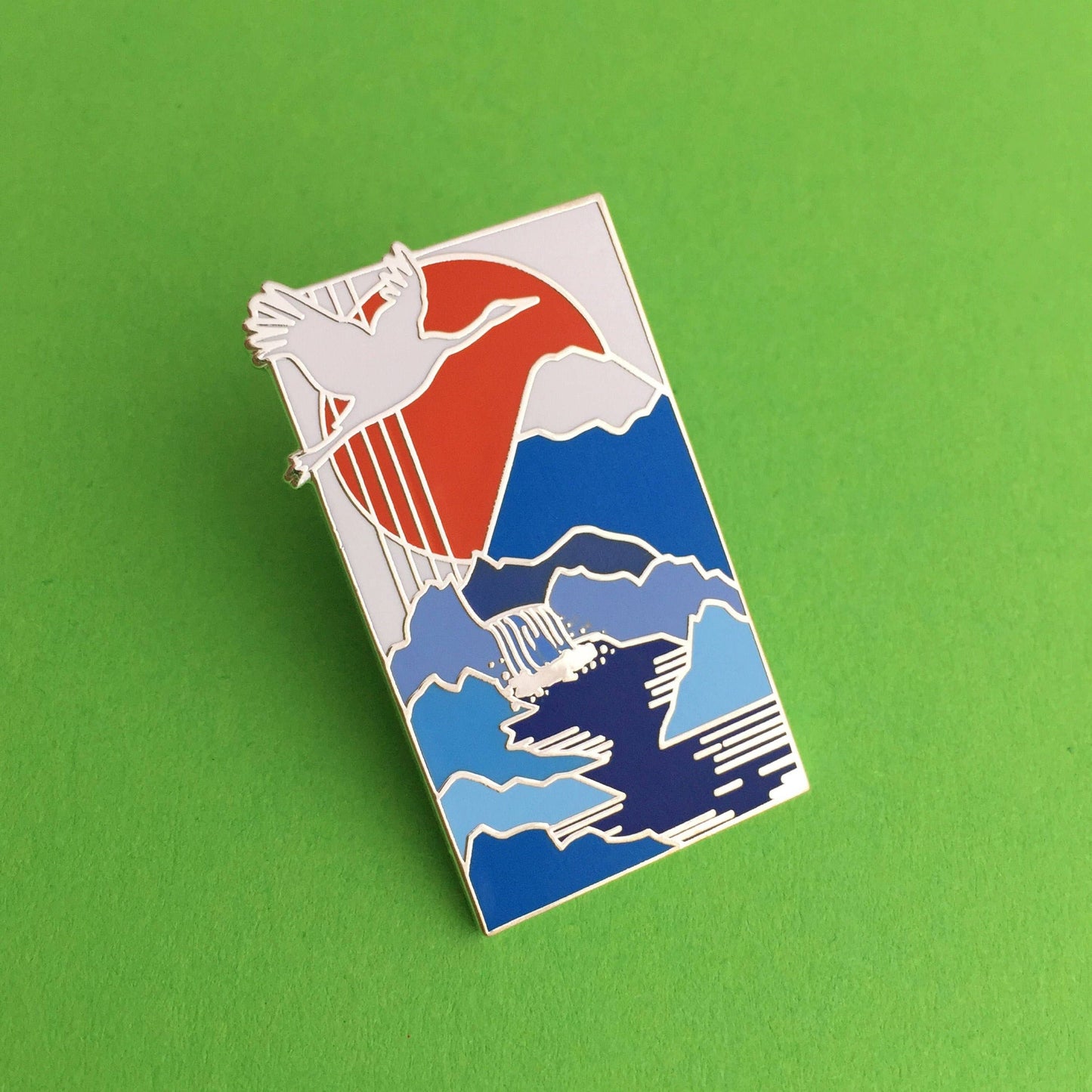 A rectangular pin, with an image of a Japanese style painting of a white crane flying across a red sun, over a snow capped mountains with a river flowing from the mountain.
Pin is on a green background.