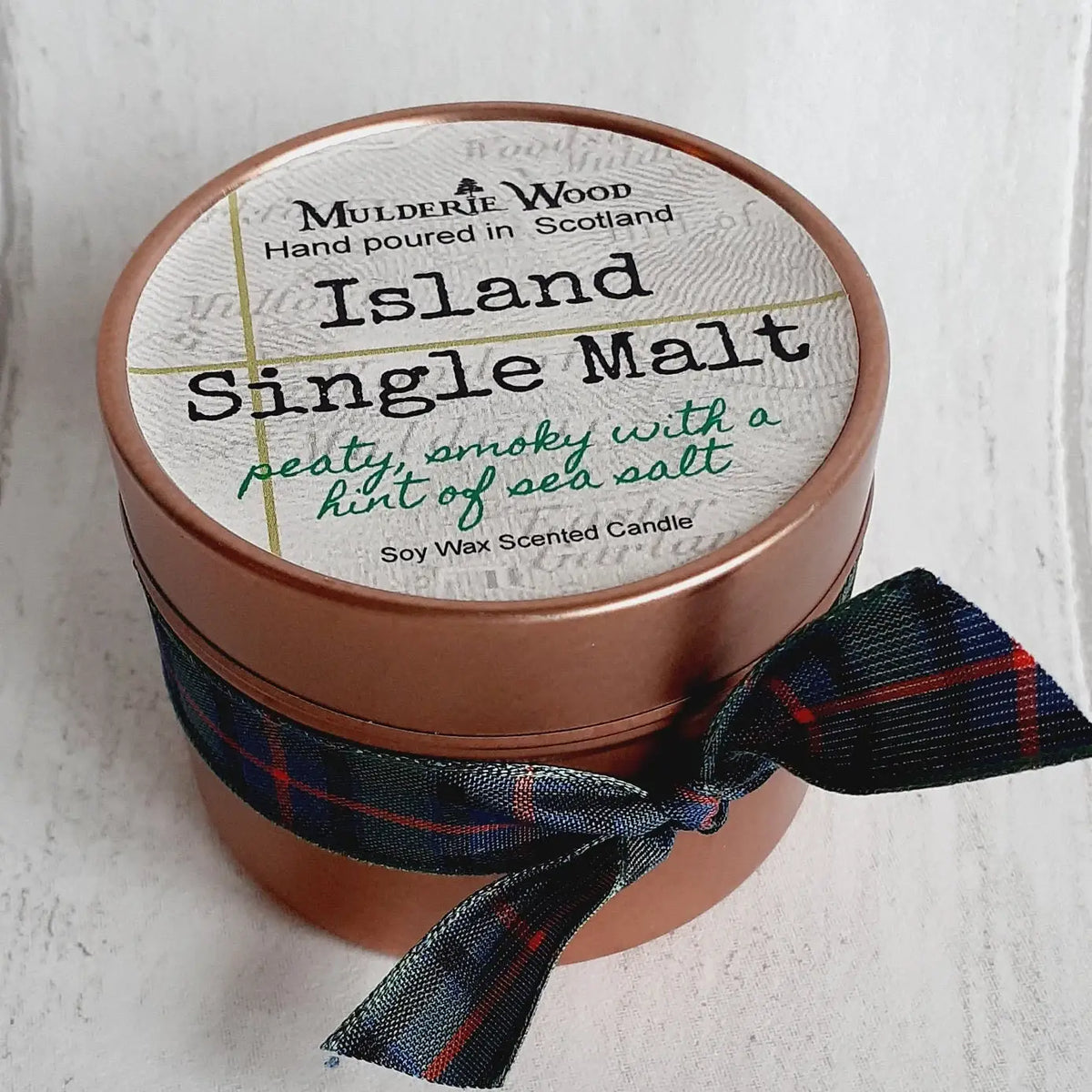 A round copper-colored candle tin with a white label reading 'Island Single Malt' by Mulderie Wood, tied with a dark plaid ribbon, on a white textured background.