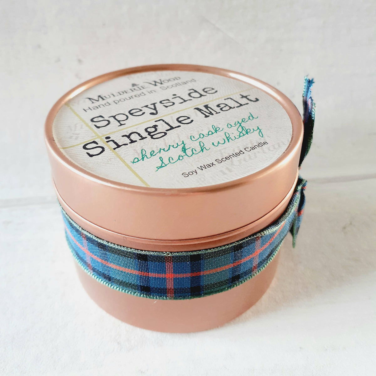 A single-wick soy wax scented candle in a round metallic tin with a copper colour finish, wrapped with a blue, green and red plaid ribbon. The lid features a cream-colored label with black and green text reading 'Speyside Single Malt'.