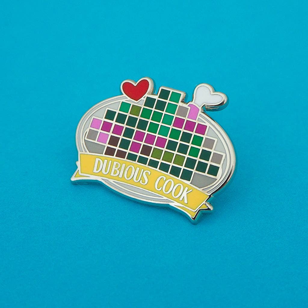 A novelty enamel pin shaped like a frying pan with a colorful pixelated grid inside, a red heart and white steam coming out, and a yellow banner reading 'DUBIOUS COOK' on a bright blue background.