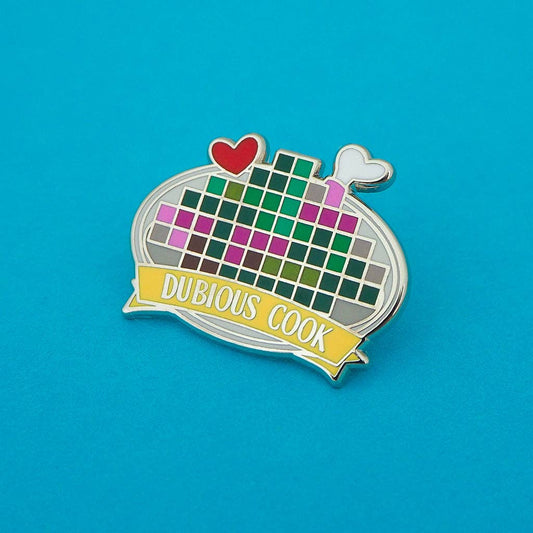 A novelty enamel pin shaped like a frying pan with a colorful pixelated grid inside, a red heart and white steam coming out, and a yellow banner reading 'DUBIOUS COOK' on a bright blue background.