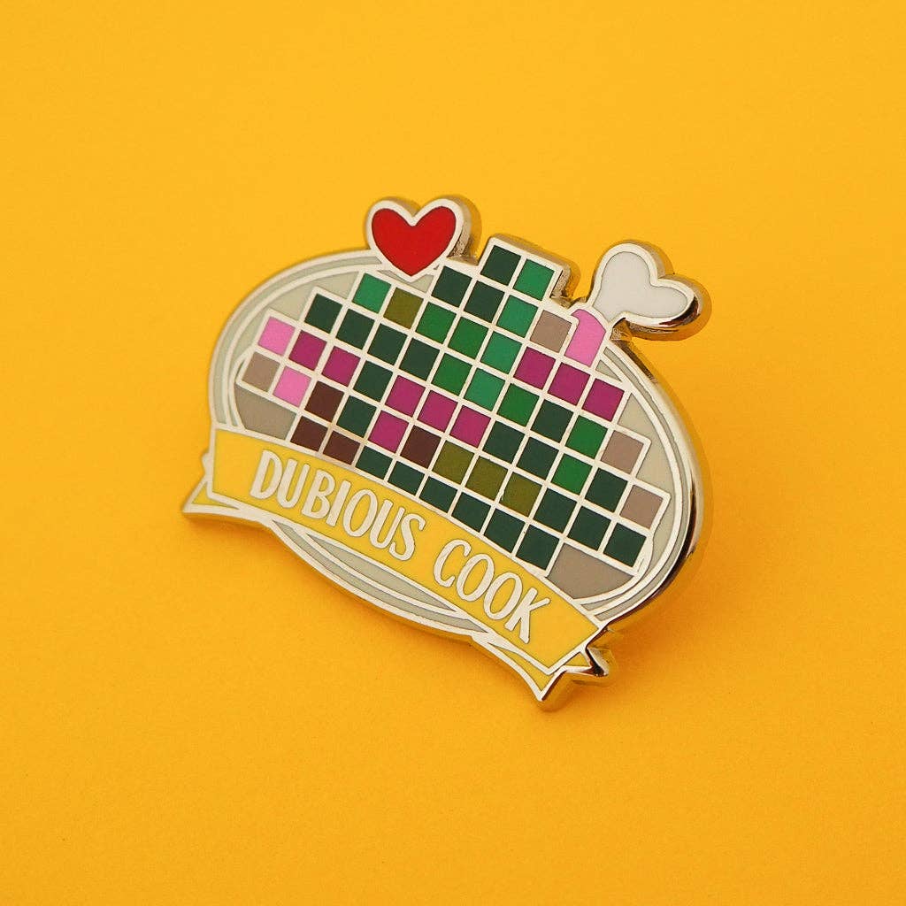 A novelty enamel pin shaped like a pan with a pixelated grid pattern and a yellow banner reading 'DUBIOUS COOK', set against a solid yellow background.