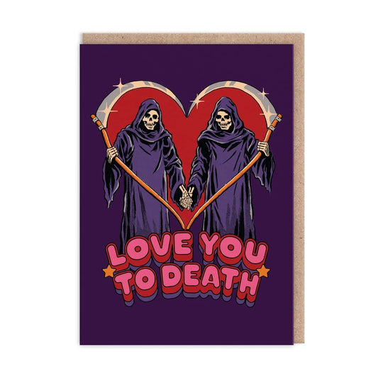 A greeting card with a kraft paper envelope, featuring a dark purple background and a graphic of two grim reapers holding scythes that form a heart shape. Below them is the text 'LOVE YOU TO DEATH' in pink and red bubble letters.