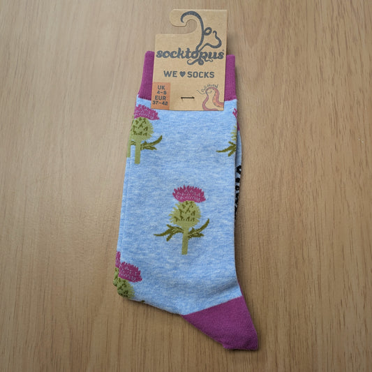 A photo of a pair of light blue socks with pink cuffs and heels. The socks feature cartoon illustrations of green thistle plants, with pink flowers on top. The cardboard hangtag is attached  to the top. Displayed on a wooden background. 