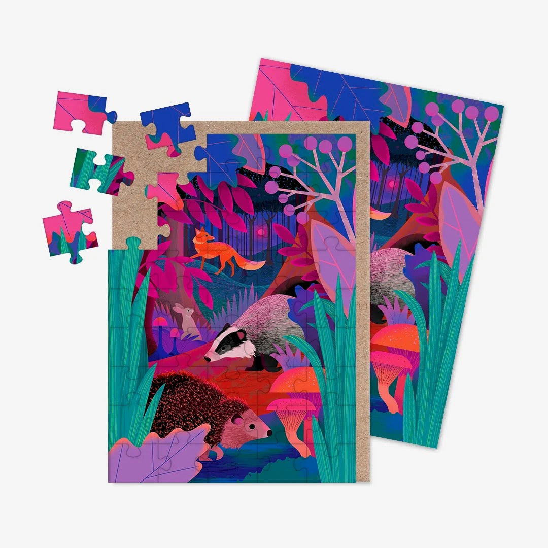 A jigsaw greeting card featuring illustrations of UK wildlife, including a badger, fox, rabbit and hedgehog in a background of shades of purple, blue, green and red with a brown kraft paper envelope visible on the left side, on a clean white background.