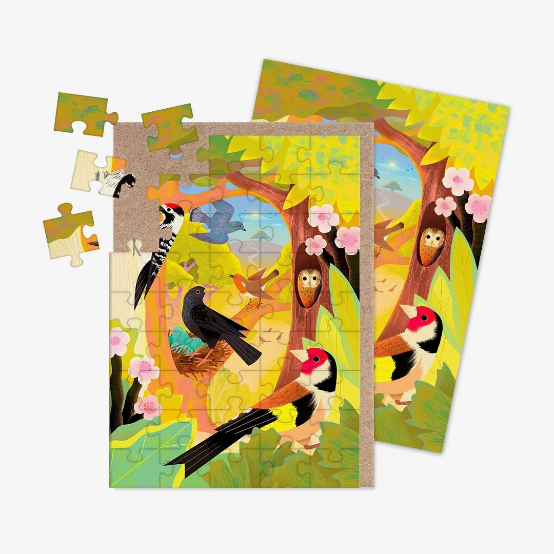A jigsaw greeting card featuring illustrations of UK birds, including a chaffinch, robin, crow and woodpecker in with a background in shades of green, yellow and brown with a brown kraft paper envelope visible on the left side, on a clean white background.