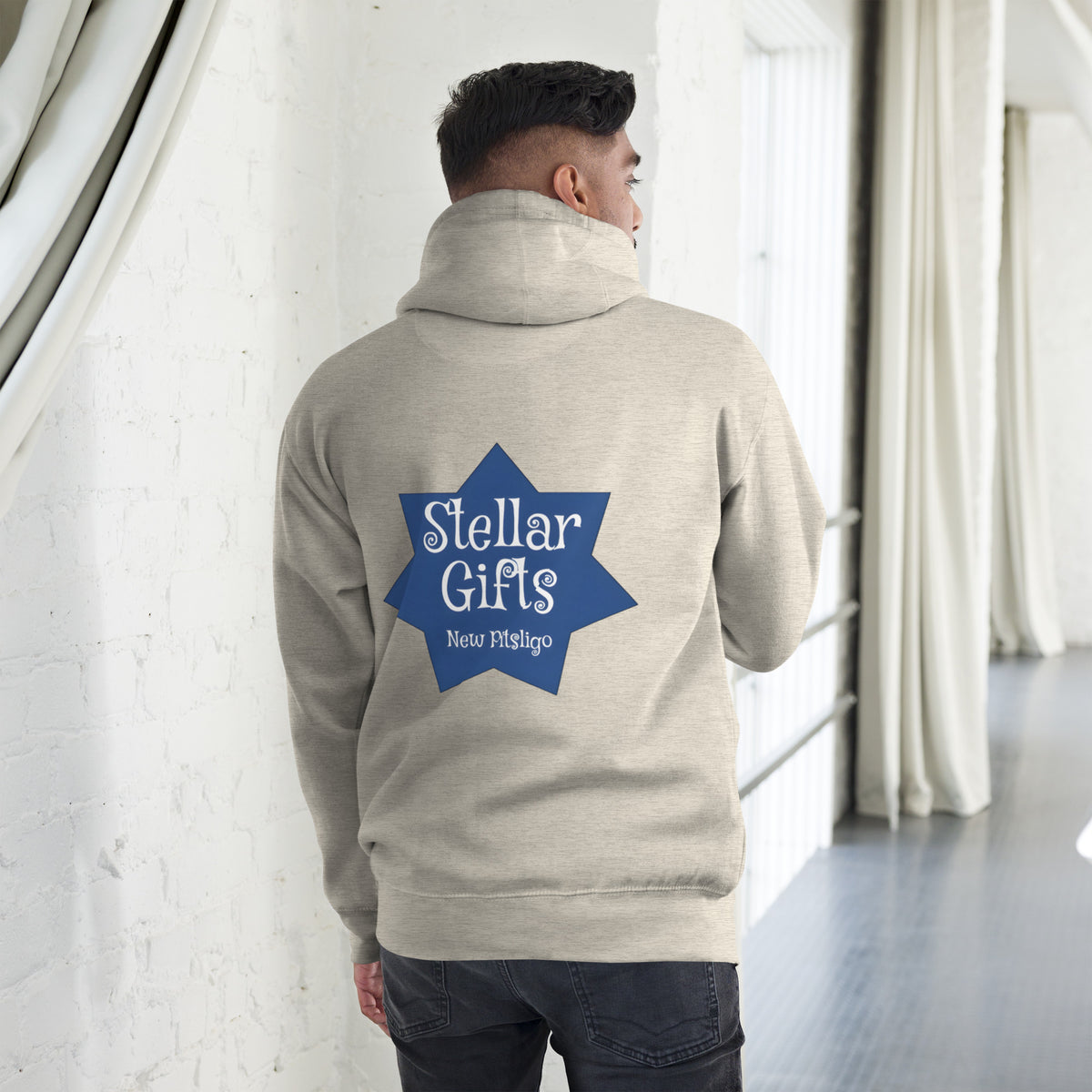 A man wearing a light brown hoodie, shot from behind on a light background. The hoodie has a dark blue, seven-sided star on the back with the STELLAR GIFTS logo written in it in white text, with "New Pitsligo" written below it.