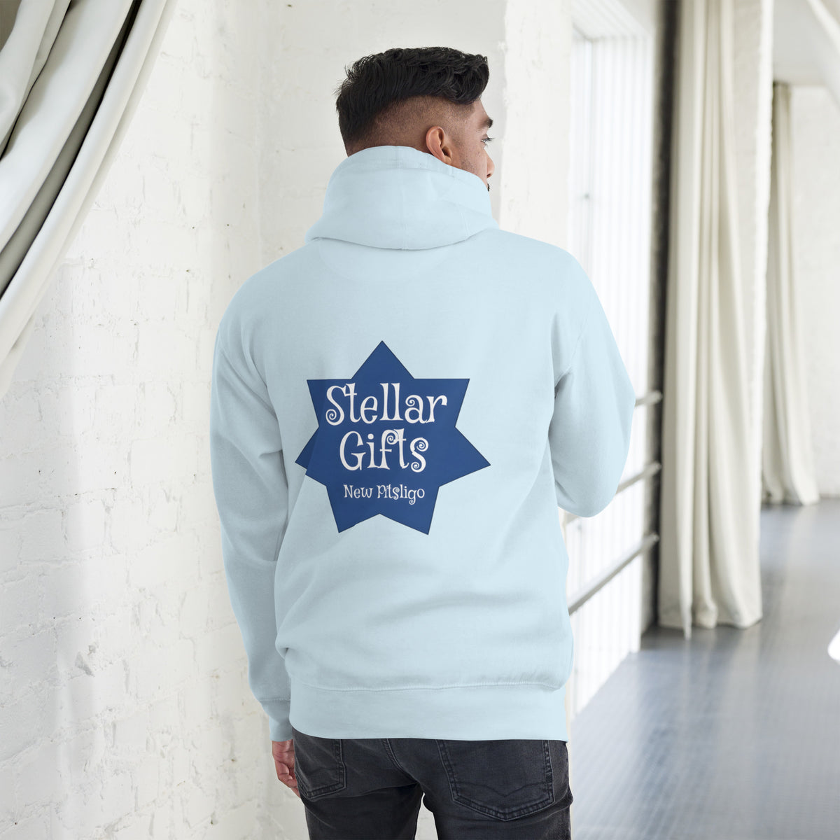 A man wearing a light blue hoodie, shot from behind on a light background. The hoodie has a dark blue, seven-sided star on the back with the STELLAR GIFTS logo written in it in white text, with "New Pitsligo" written below it.