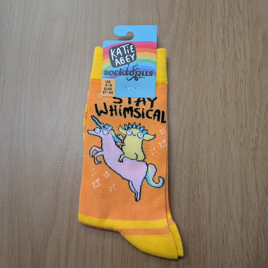 A photo of a pair of orange socks with yellow cuffs and heels. The socks feature an illustration of a yellow cartoon figure riding a pink unicorn, with the text "STAY WHIMSICAL" in black text above. The cardboard hangtag is attached  to the top. Displayed on a wooden background. 