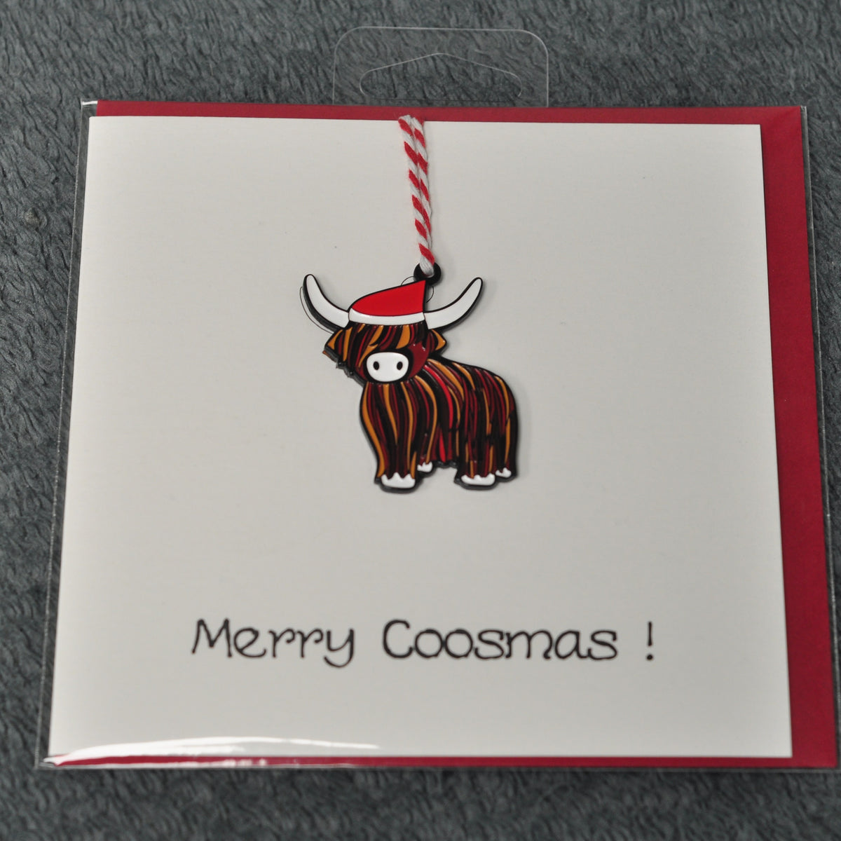 A Scottish Christmas greeting card featuring a cartoon Highland cow ornament with a red Santa hat, on a red envelope with 'Merry Coosmas!' written in black script.