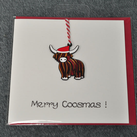 A Scottish Christmas greeting card featuring a cartoon Highland cow ornament with a red Santa hat, on a red envelope with 'Merry Coosmas!' written in black script.