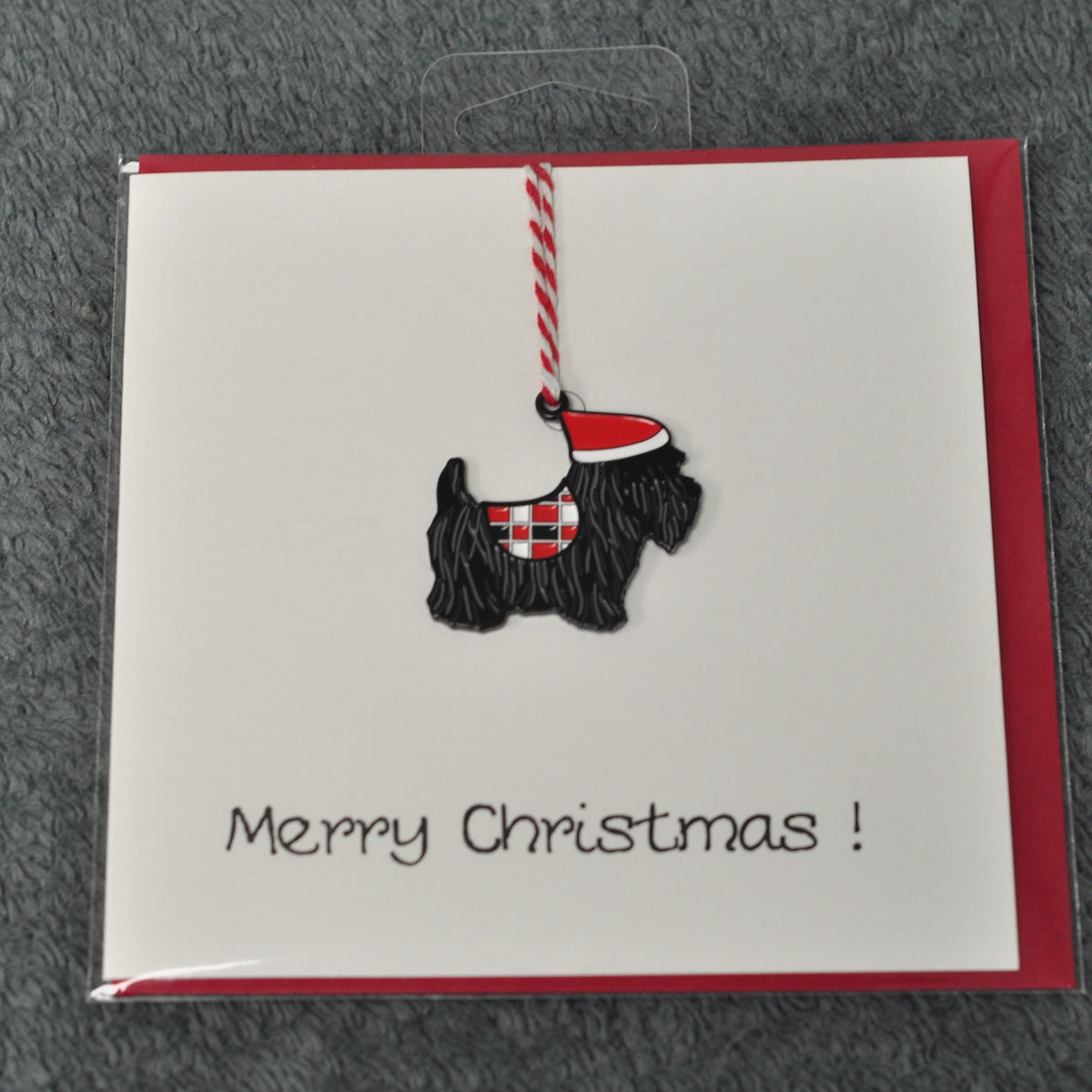 A Christmas greeting card with a red envelope and a black-and-white Scottie dog ornament hanging from a red-and-white striped string, with the text 'Merry Christmas!' on a white textured background.